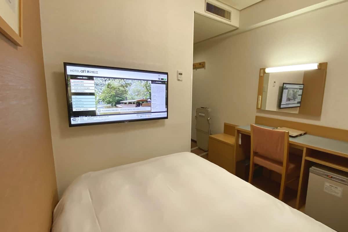 Ein Hotelzimmer mit einem Bett, einem Schreibtisch, einem Stuhl, einem kleinen Kühlschrank und einem an der Wand montierten Fernseher, der eine Hotelwebseite anzeigt.