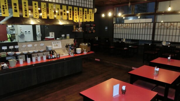Interno di un ristorante con un bancone che espone diversi piatti, tavoli con menu e un'estetica tradizionale giapponese.