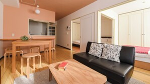 A modern living room with a black leather sofa, wooden dining table, and chairs. There is a kitchen area with a countertop and cabinets. The room has a large window providing natural light. - Dai37 Matsui Building (Sapporo)