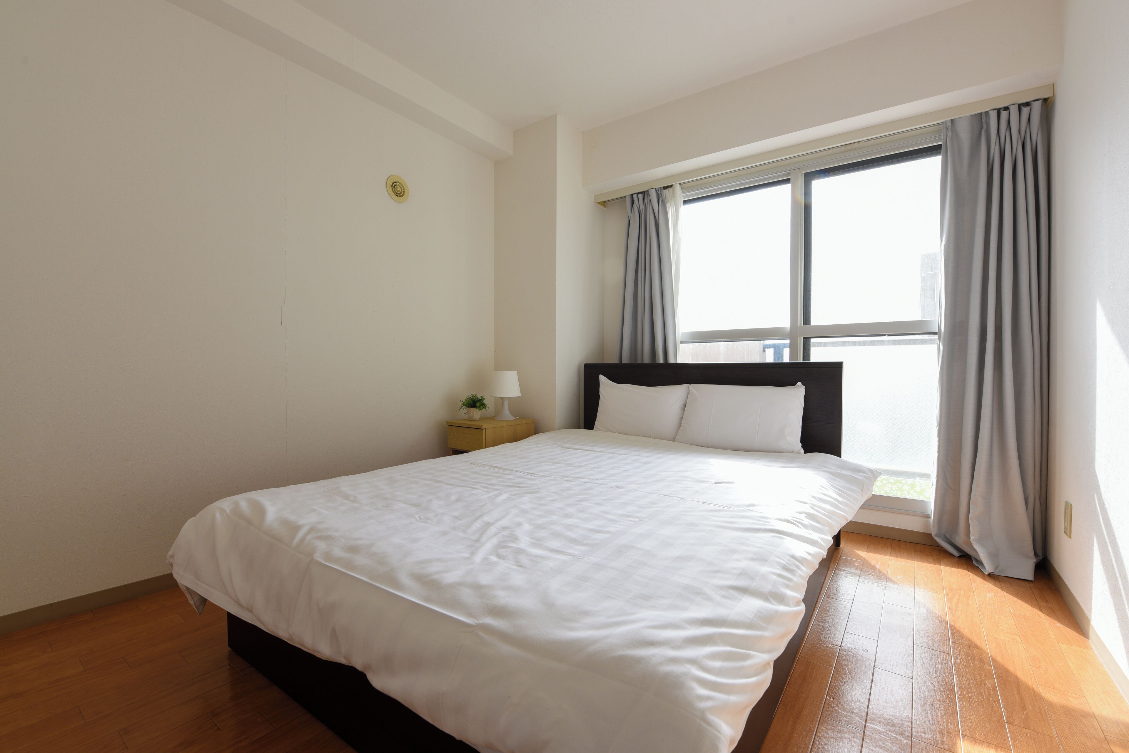 A bedroom with a bed, a nightstand with a lamp, a window with curtains, and a wooden floor.