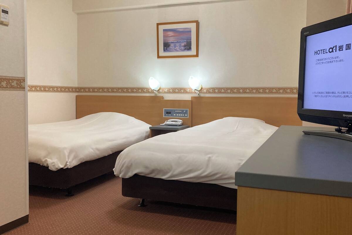 A hotel room with two beds, a desk, a television, and a wall-mounted thermostat.