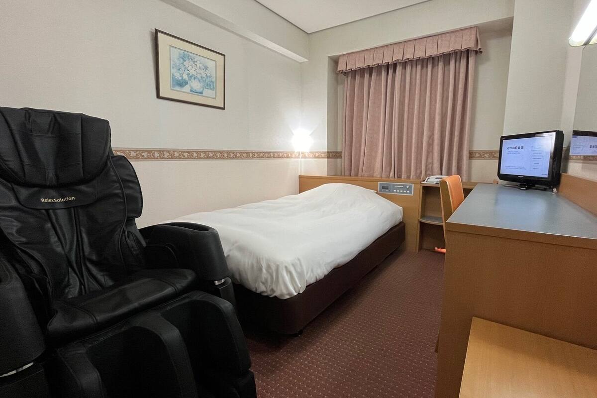 A hotel room with a bed, a massage chair, a desk with a computer, and a window with curtains.