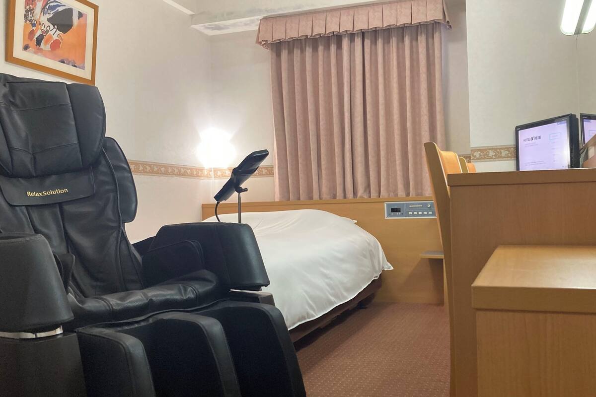 A hotel room with a massage chair, a bed with a white comforter, a bedside table, a television, and a window with curtains.
