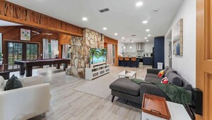 A modern living room with a pool table, a sofa, a television, and a kitchen area in the background. - The Cheswick A I Team Joseph Ellen (Orange Park)