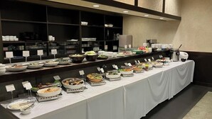 Tarjolla on runsas buffet, jossa on erilaisia ruokia ja astioita, kuten kulhoja ja tarjottimia, ja ne on aseteltu pitkälle pöydälle, jonka päällä on valkoinen pöytäliina.