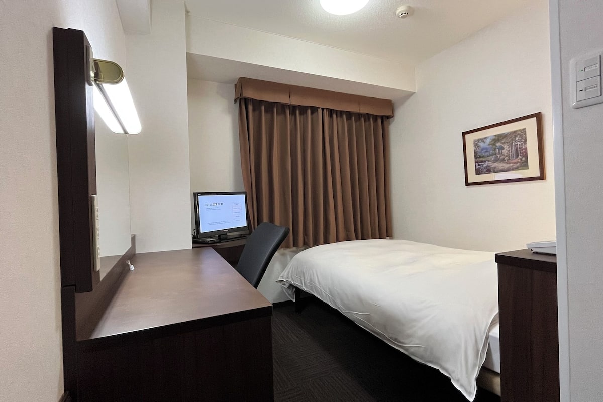 A hotel room with a bed, a desk with a computer, a chair, a window with curtains, and a framed picture on the wall.