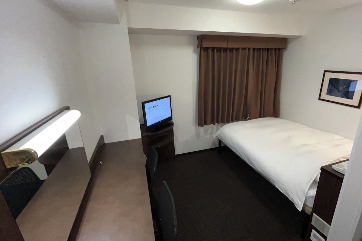 A hotel room with a bed, a desk with a computer, a chair, a lamp, and a window with curtains.