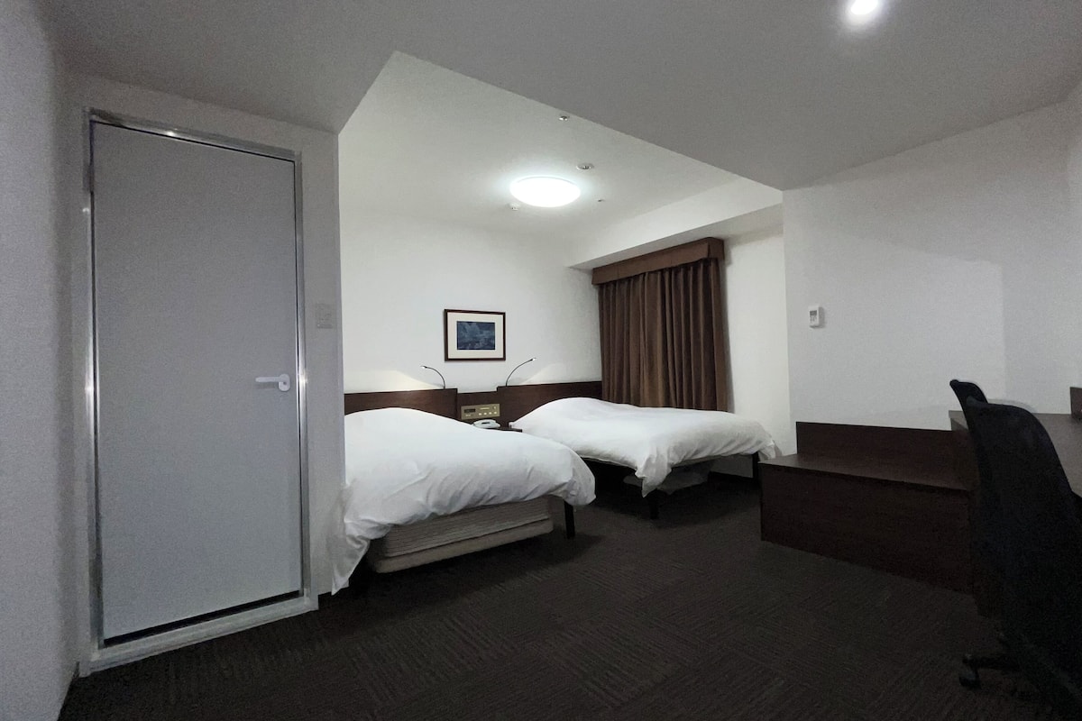 A hotel room with two single beds, a desk, a chair, a wardrobe, and a ceiling light.
