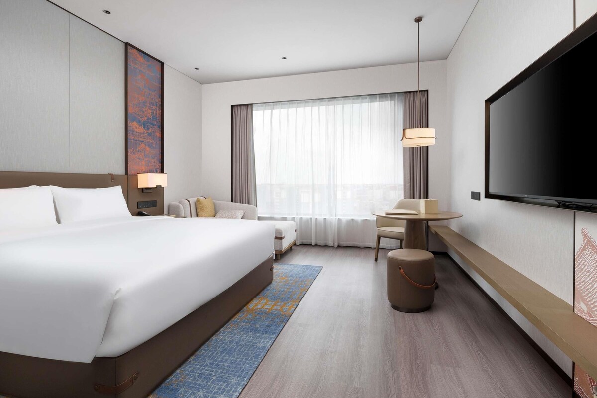 Ein modernes Hotelzimmer mit einem großen Bett, einem Schreibtisch, einem Fernseher und einem großen Fenster mit Vorhängen.
