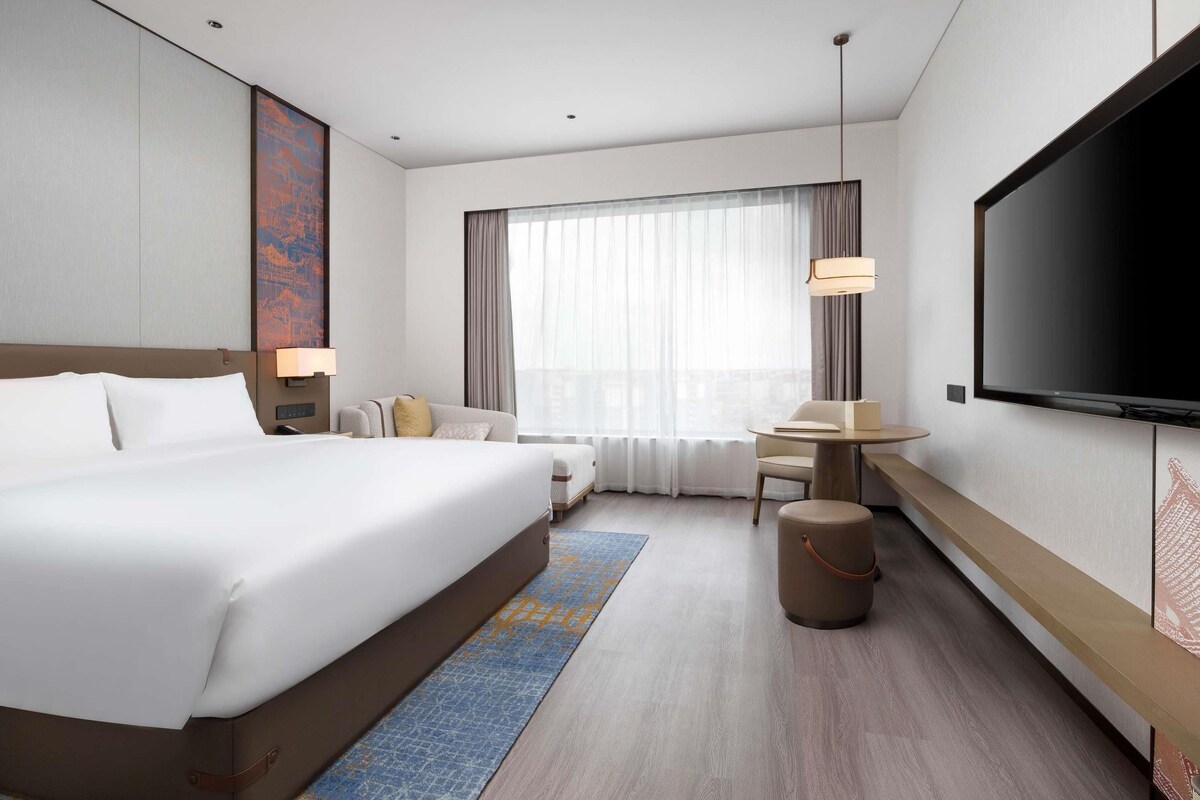 Ein Hotelzimmer mit einem großen Bett, einem Schreibtisch, einem Sessel, einem Fernseher und einem Fenster mit Vorhängen.