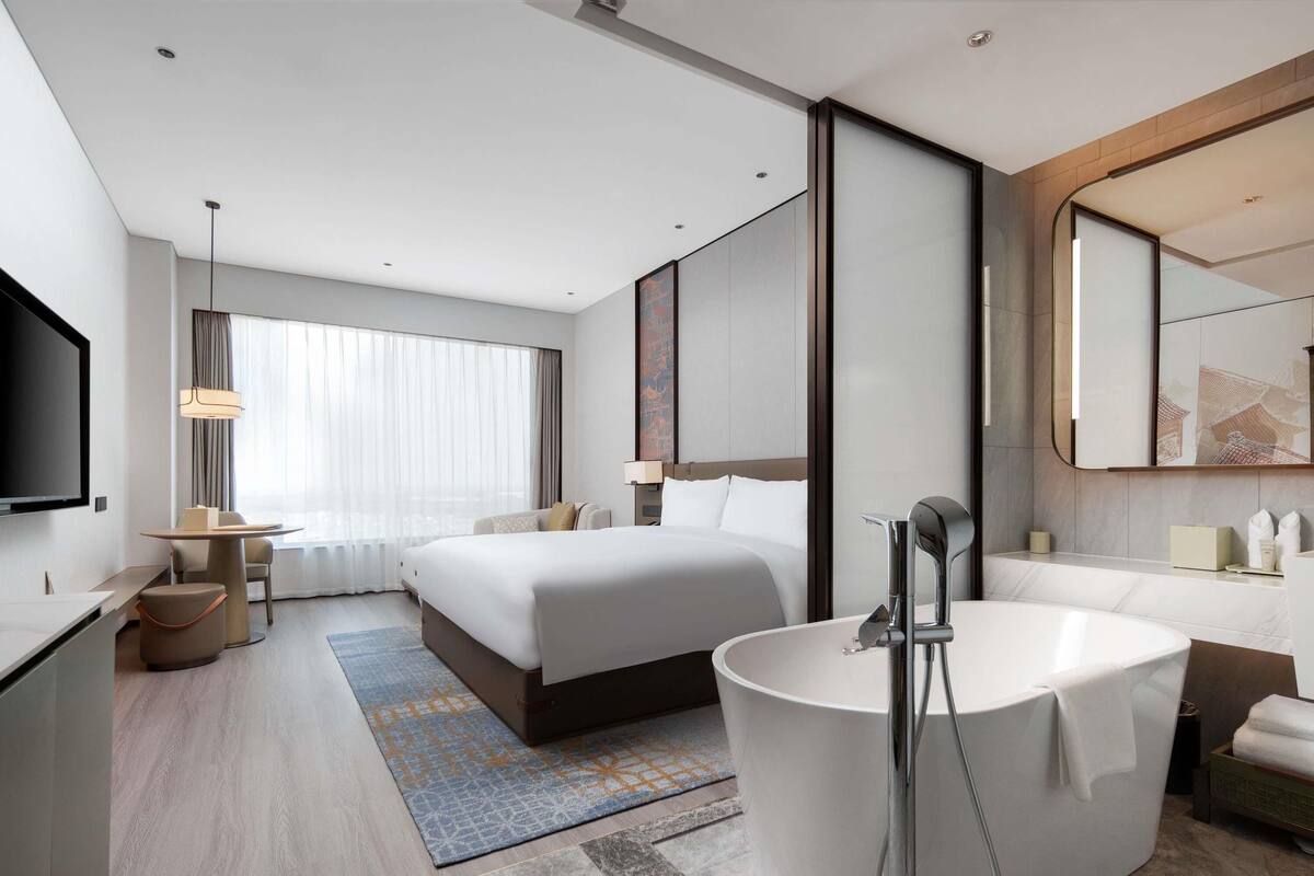 Een moderne hotelkamer met een groot bed, een aparte badkamer met bad, een bureau met stoel en een grote spiegel.
