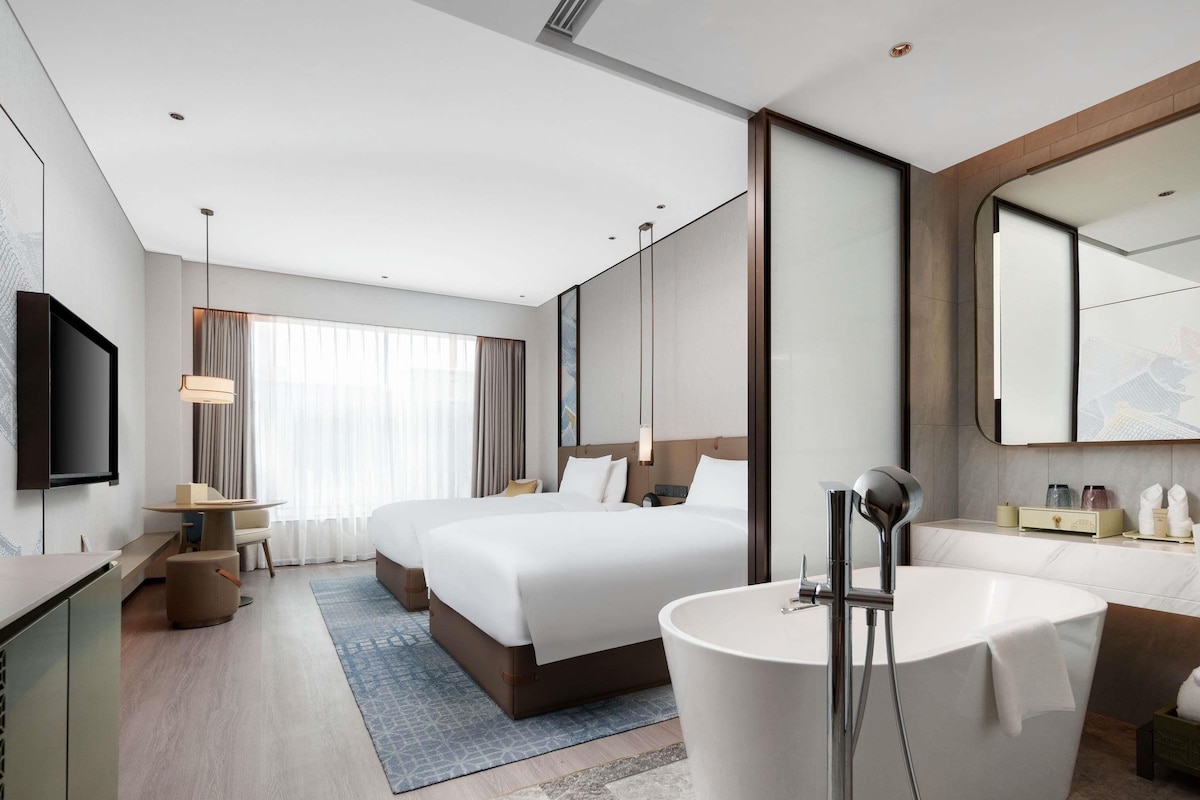 A modern hotel room with a large bed, a separate sitting area, a bathtub, and a flat-screen TV.