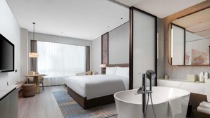 Habitación de hotel moderna con una cama grande, un baño separado con bañera, un escritorio con silla y un espejo amplio.