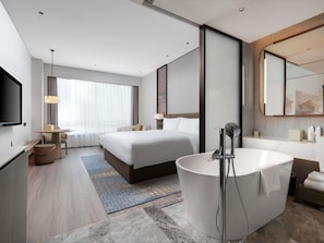 A modern hotel room with a large bed, a separate bathroom with a bathtub, a desk with a chair, and a large mirror.