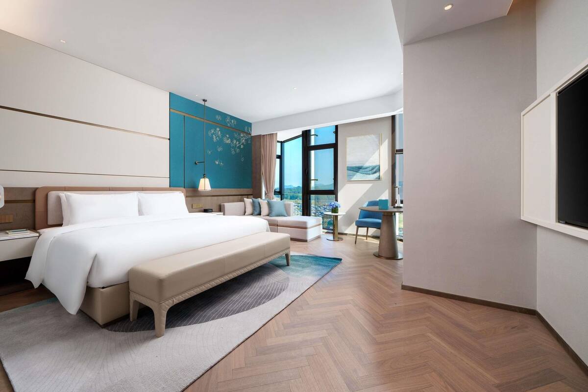 Una habitación de hotel moderna con una cama grande, una zona de estar, una mesita y un amplio ventanal con vistas a la ciudad.