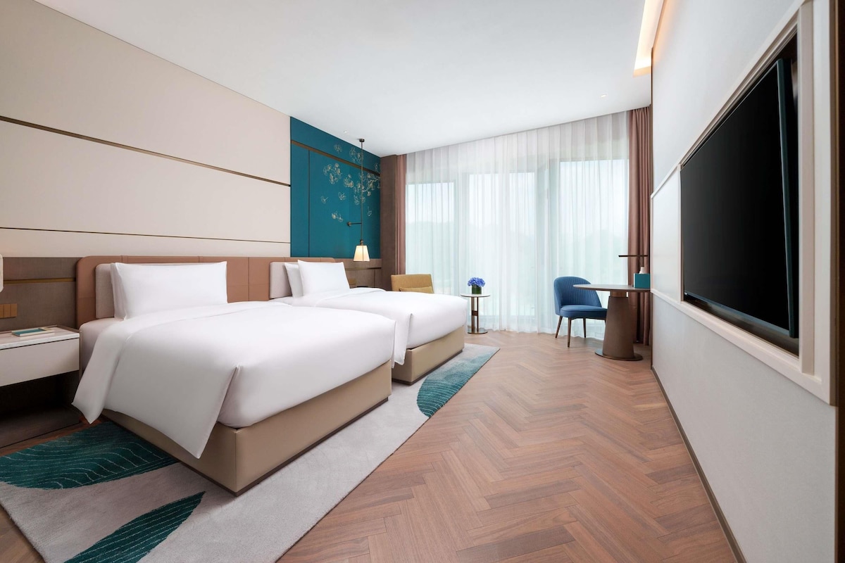 A modern hotel room with two beds, a desk, a chair, and a large TV screen.