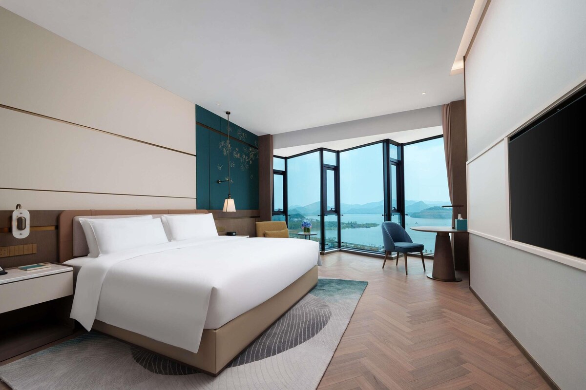 A modern hotel room with a large bed, a desk, a chair, and a large window offering a view of a lake and mountains.