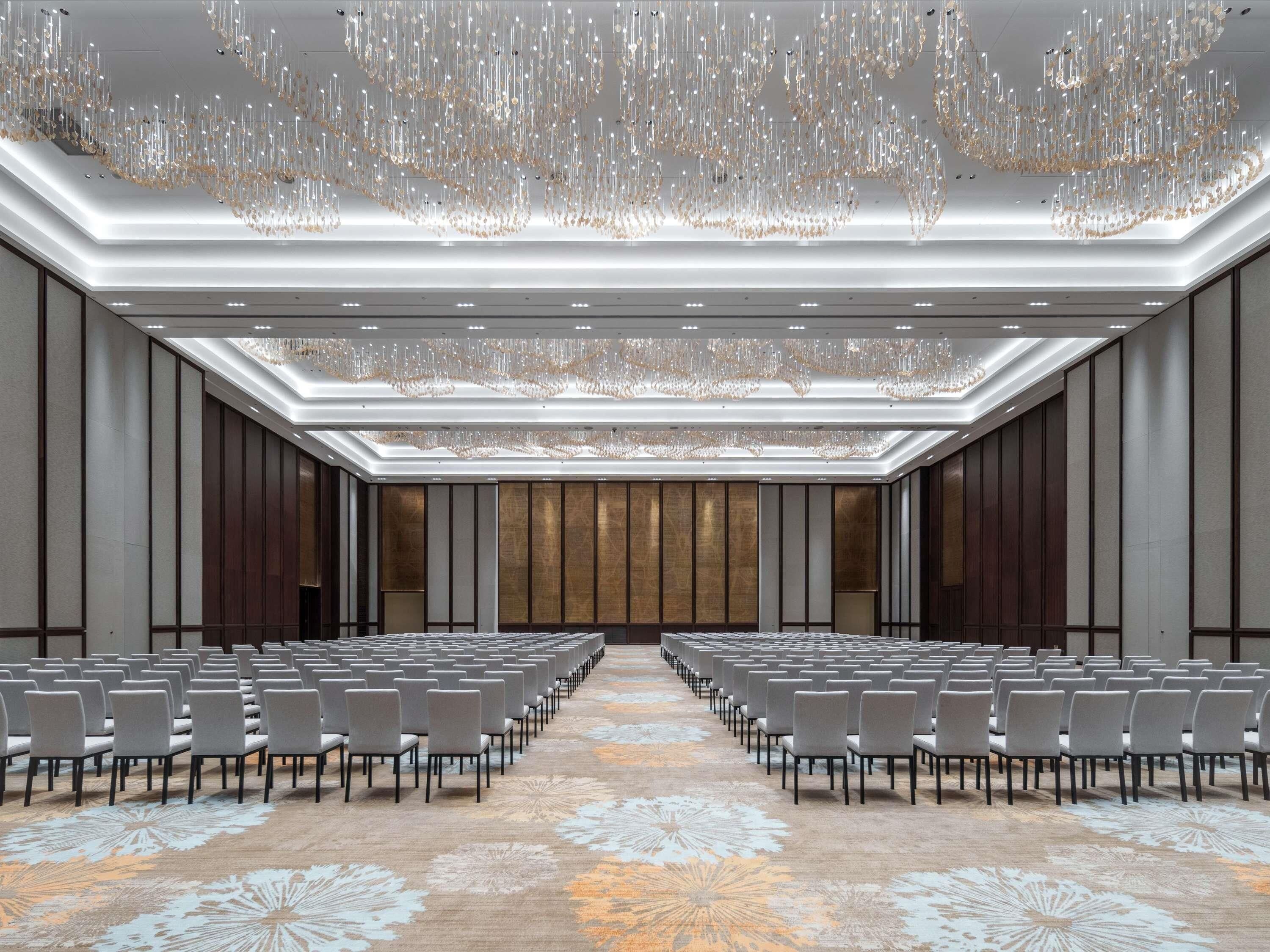 Un salón de conferencias con filas de sillas, alfombra estampada y un techo con candelabros colgantes.