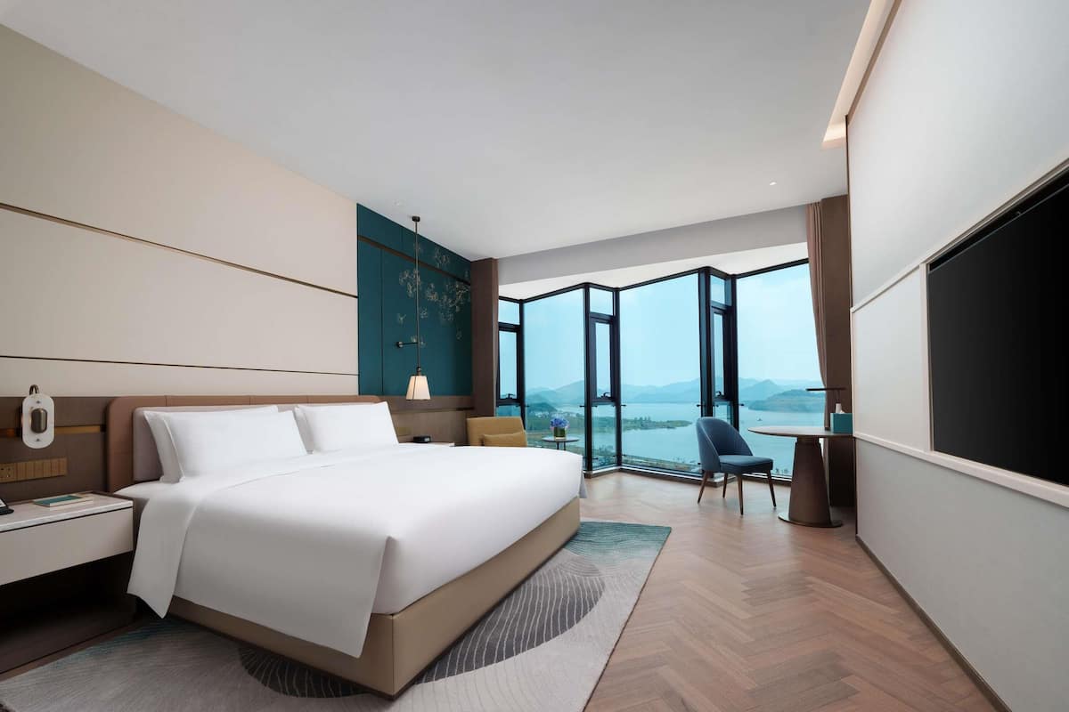 Una habitación de hotel moderna con una cama grande, un escritorio, una silla y un amplio ventanal con vistas a un lago y montañas.