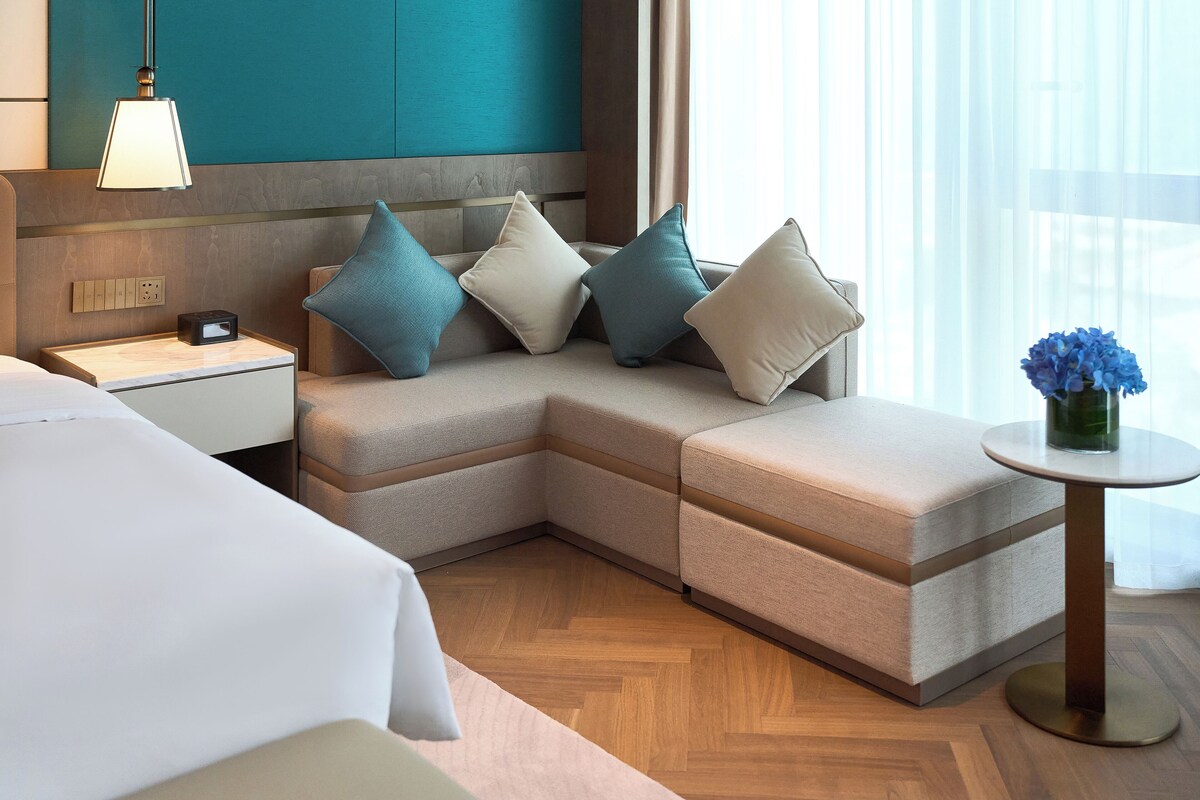 A modern hotel room with a sectional sofa, a bedside table with a lamp, a small round table with a vase, and a wooden floor.