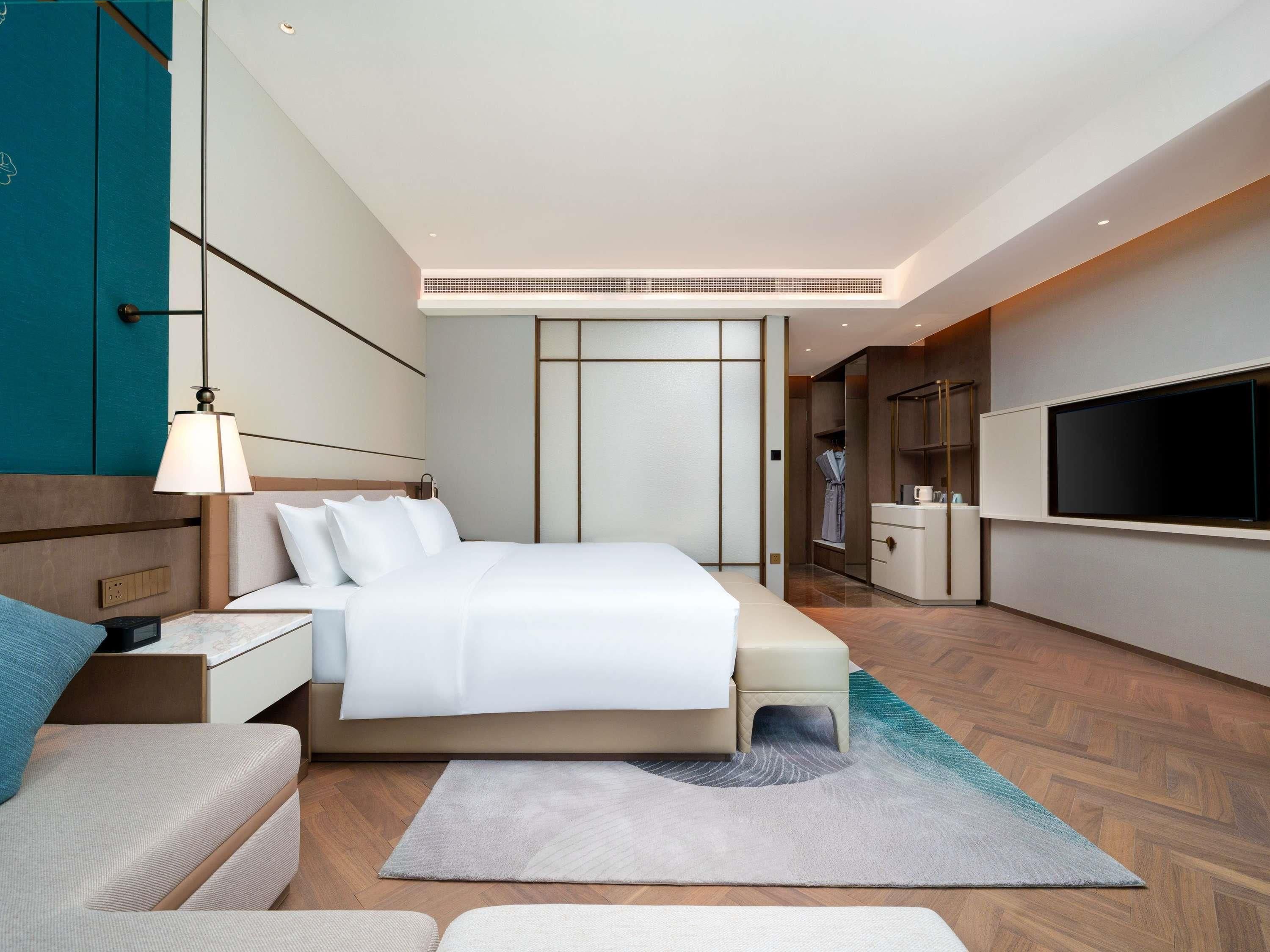 A modern hotel room with a bed, bedside table, lamp, TV, wardrobe, and a small seating area.