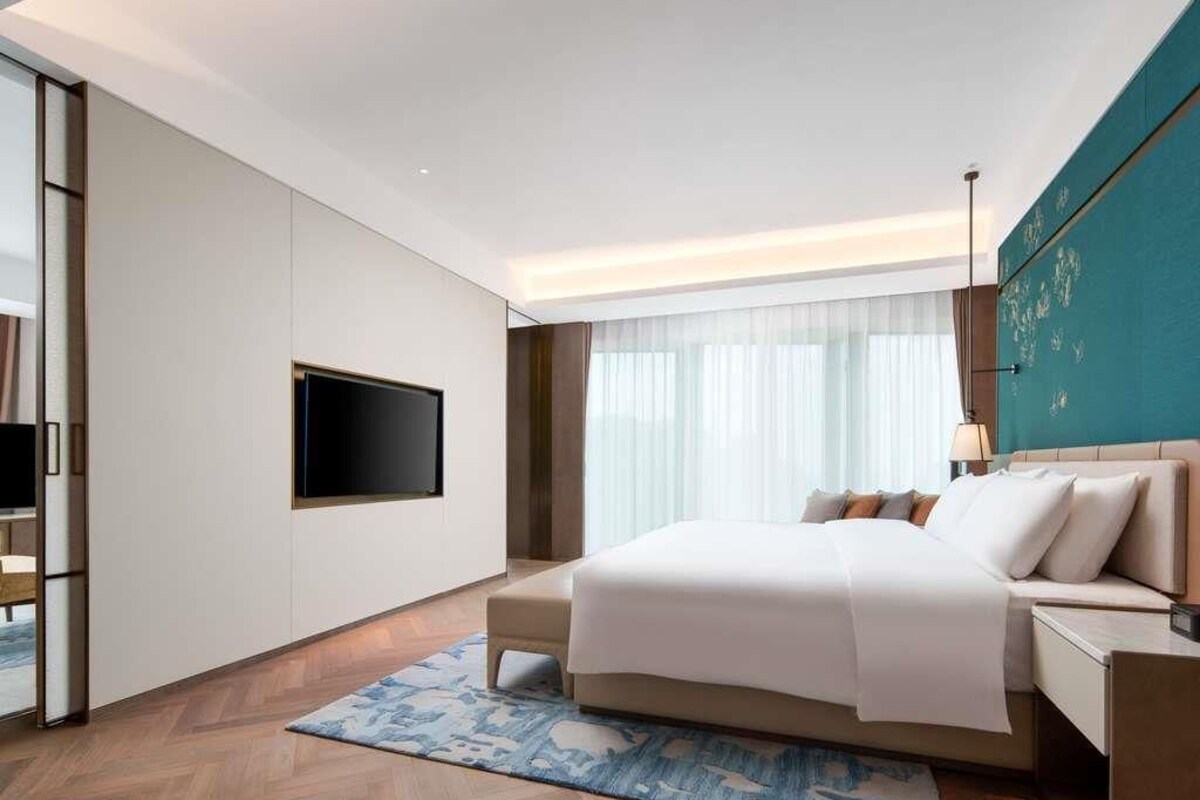 A modern hotel room with a large bed, a flat-screen TV, a desk, and a window with sheer curtains.