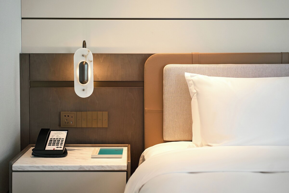 A hotel room with a bed, a bedside table with a telephone, a notepad, and a wall-mounted light fixture.