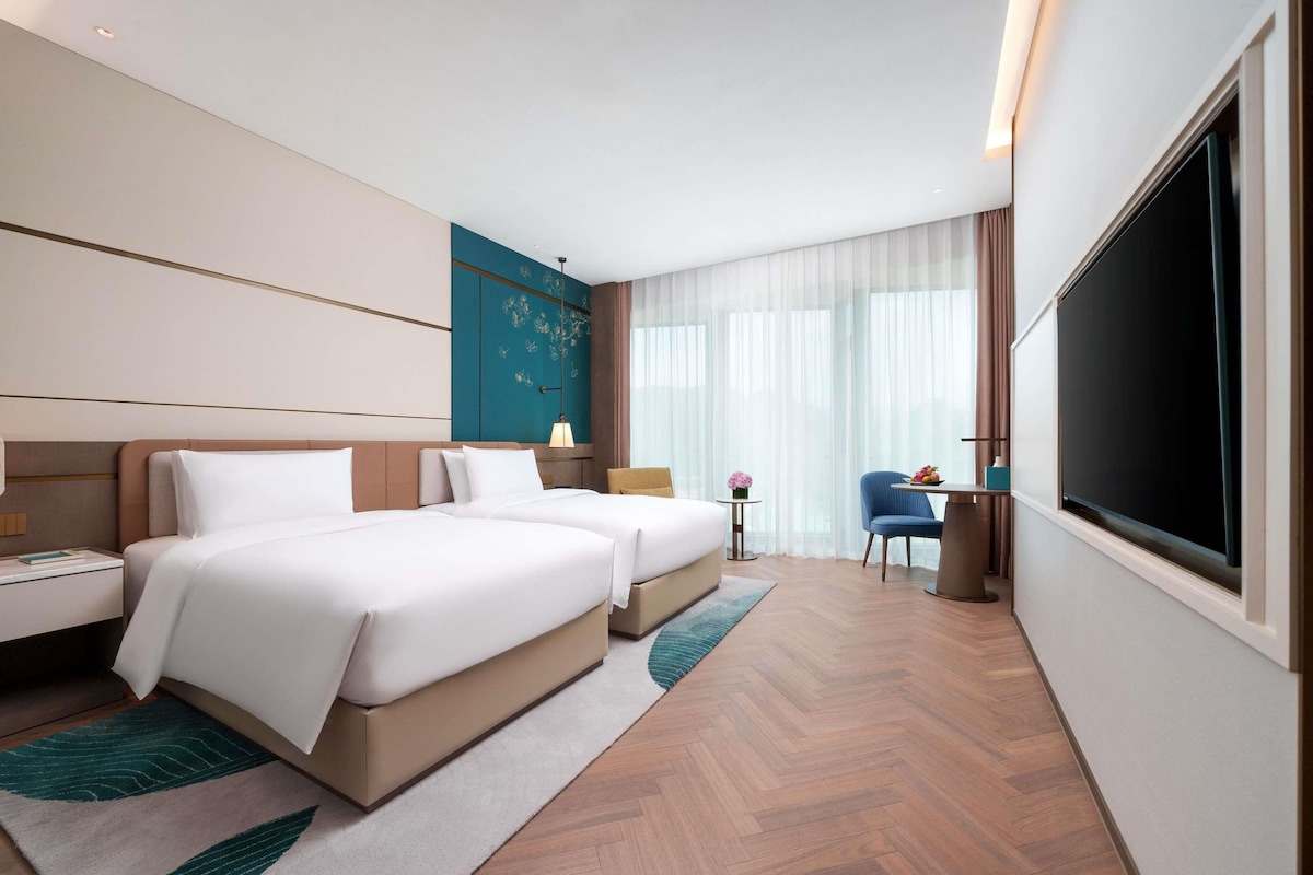 A hotel room with two beds, a desk, a chair, a TV, and a large window with curtains.