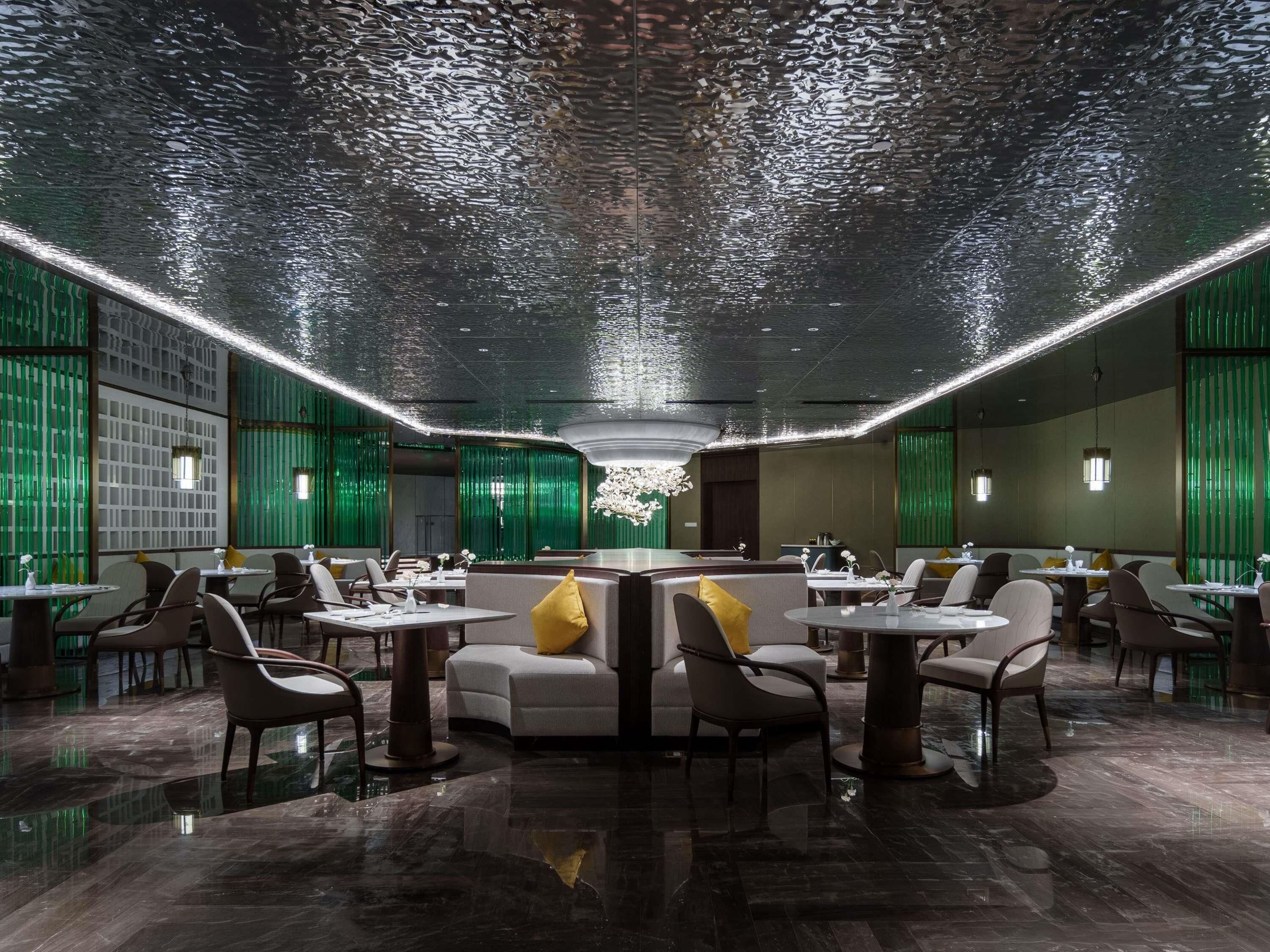 Een modern restaurant met een opvallend plafondontwerp, groene wanden en een centraal zitgedeelte met ronde tafels en stoelen.