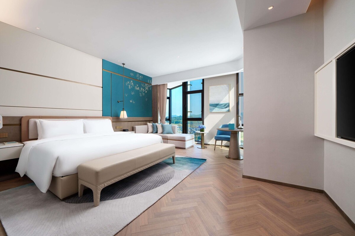 A modern hotel room with a large bed, a seating area, a small table, and a large window offering a city view.