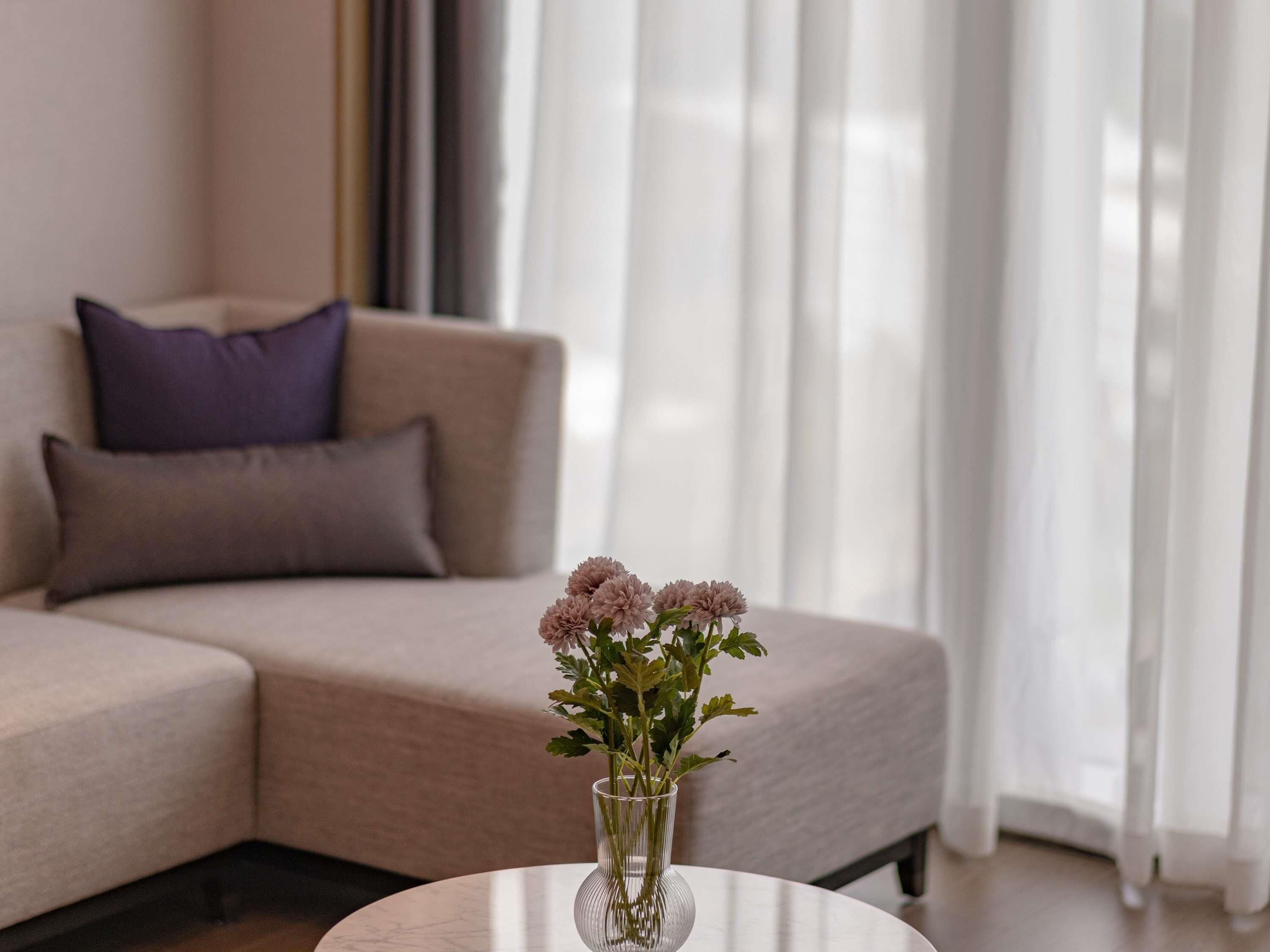 A living room with a beige sofa, a glass vase with flowers on a round table, and sheer curtains.