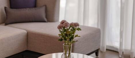 A living room with a beige sofa, a glass vase with flowers on a round table, and sheer curtains.
