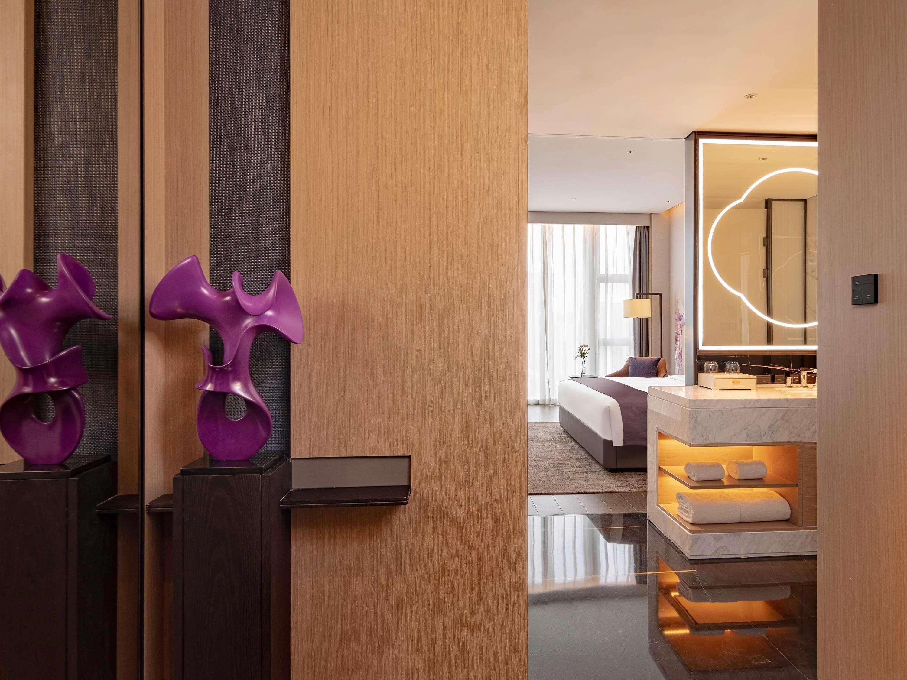 Una habitación de hotel moderna con una puerta de madera, un espejo grande con luz circular, una cama con cabecera blanca y un tocador de baño de mármol.