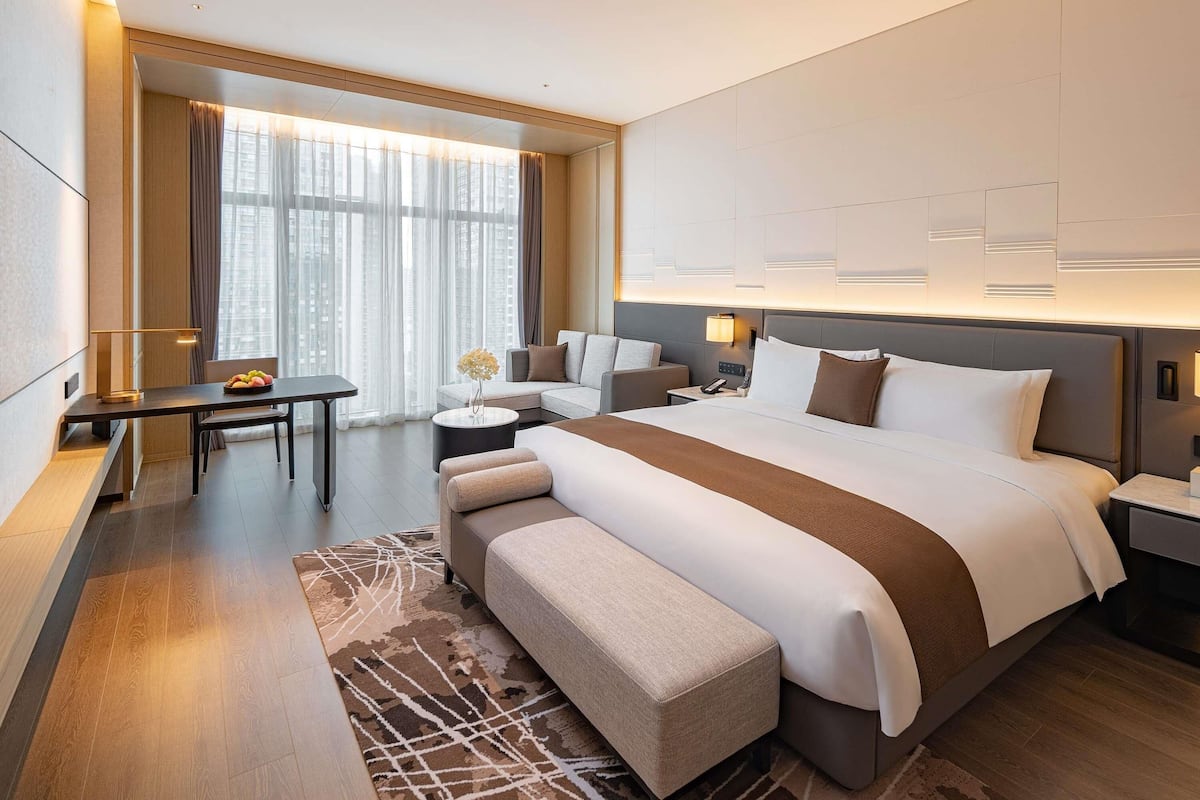 A modern hotel room with a large bed, a desk, a chair, and a view of the city.