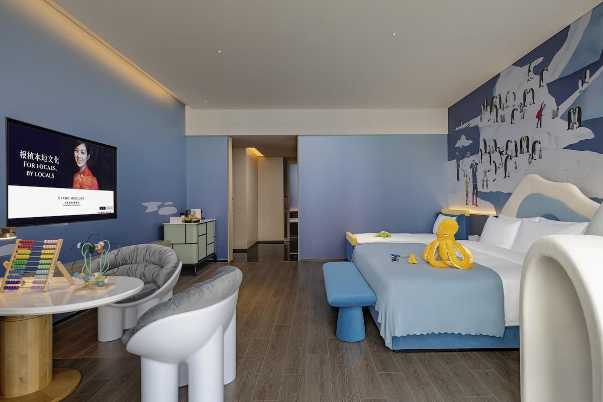 A modern hotel room with a blue and white color scheme, a large bed, a round table with an abacus, a gray armchair, and a wall-mounted TV displaying a movie poster.