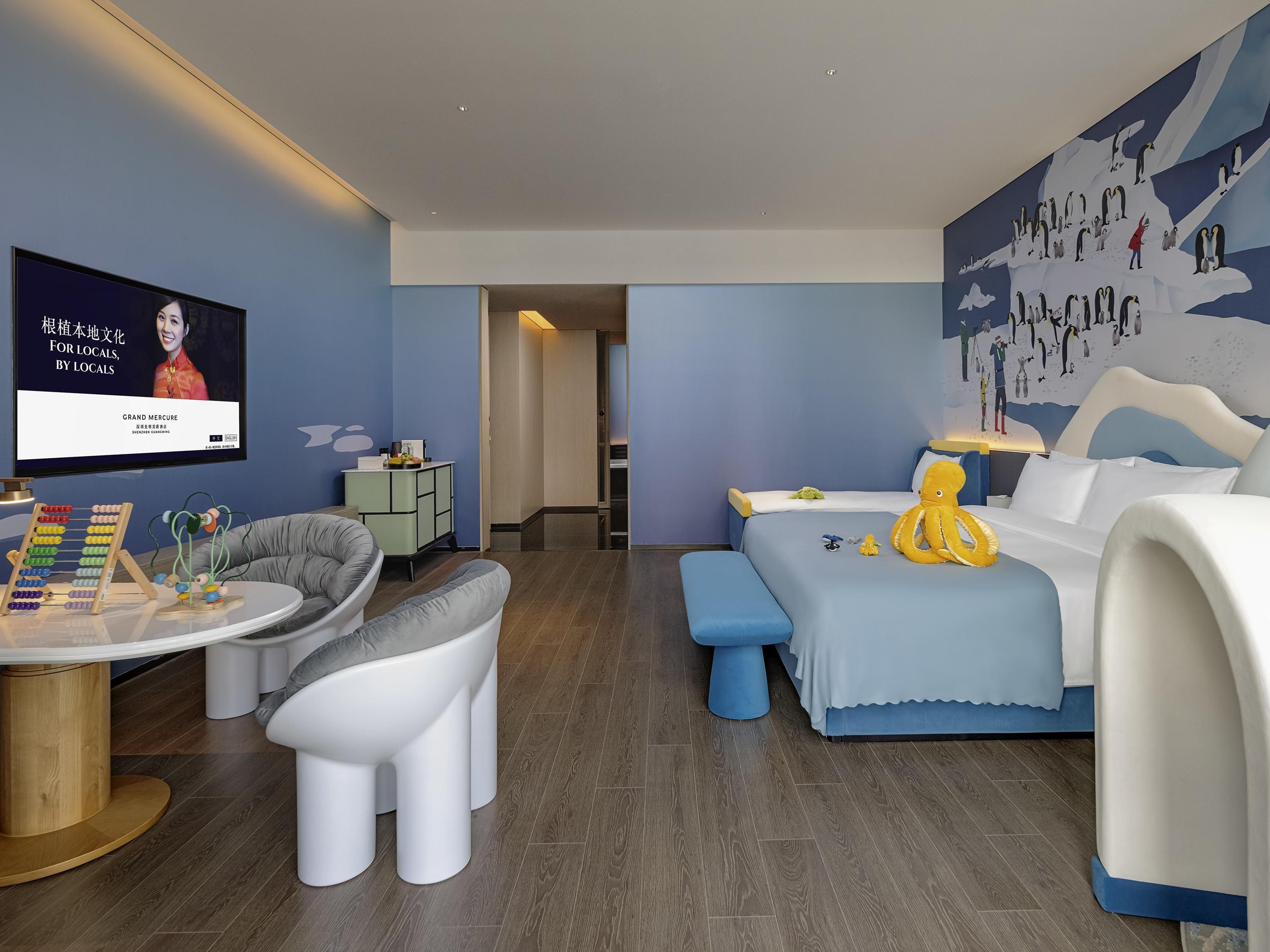 A modern hotel room with a large bed, a seating area with a round table, and a wall-mounted TV.
