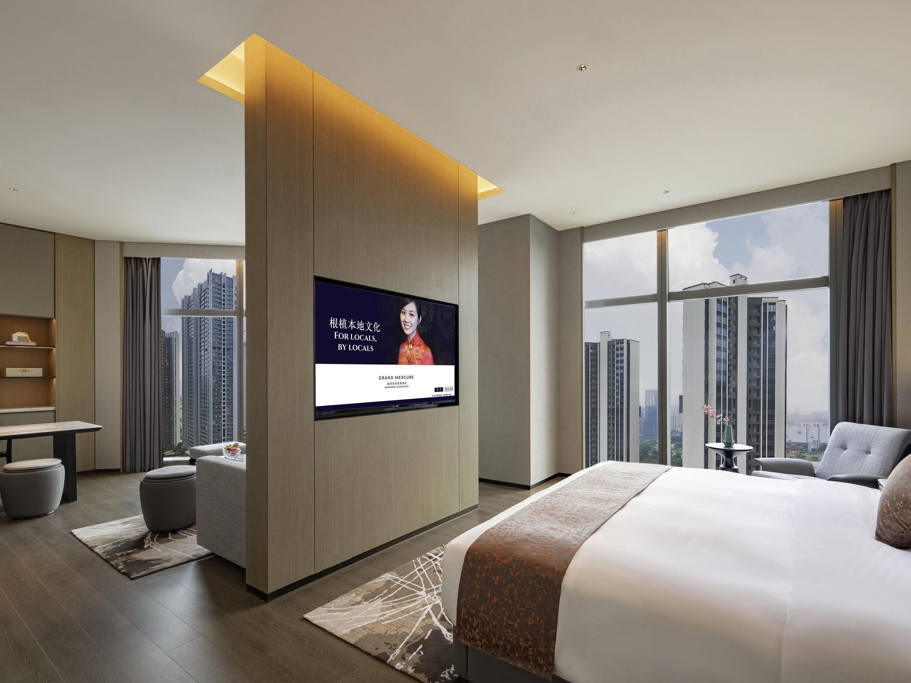 A modern hotel room with a large bed, a flat-screen TV mounted on a wooden panel, a desk, and a view of tall buildings.