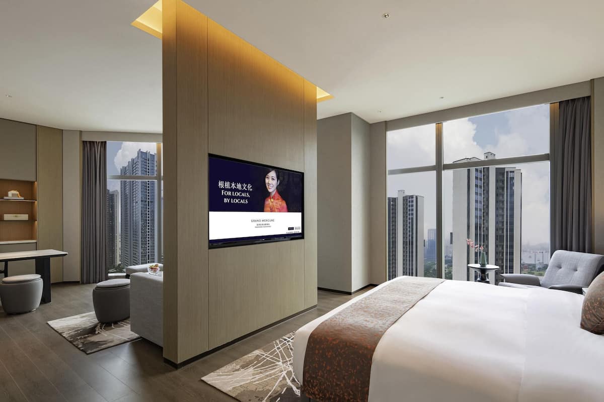 A modern hotel room with a large bed, a flat-screen TV mounted on a wooden panel, a desk, and a view of tall buildings.