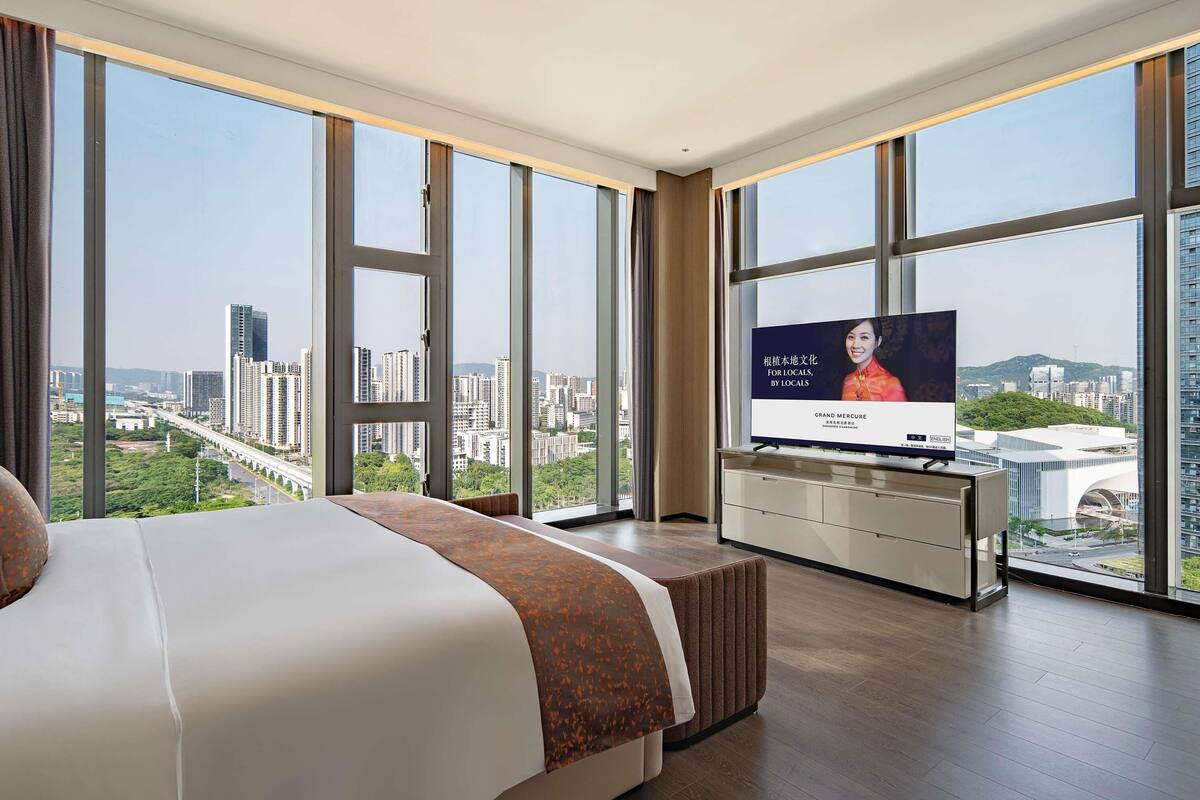 A hotel room with a large bed, a TV on a stand, and a city view through large windows.