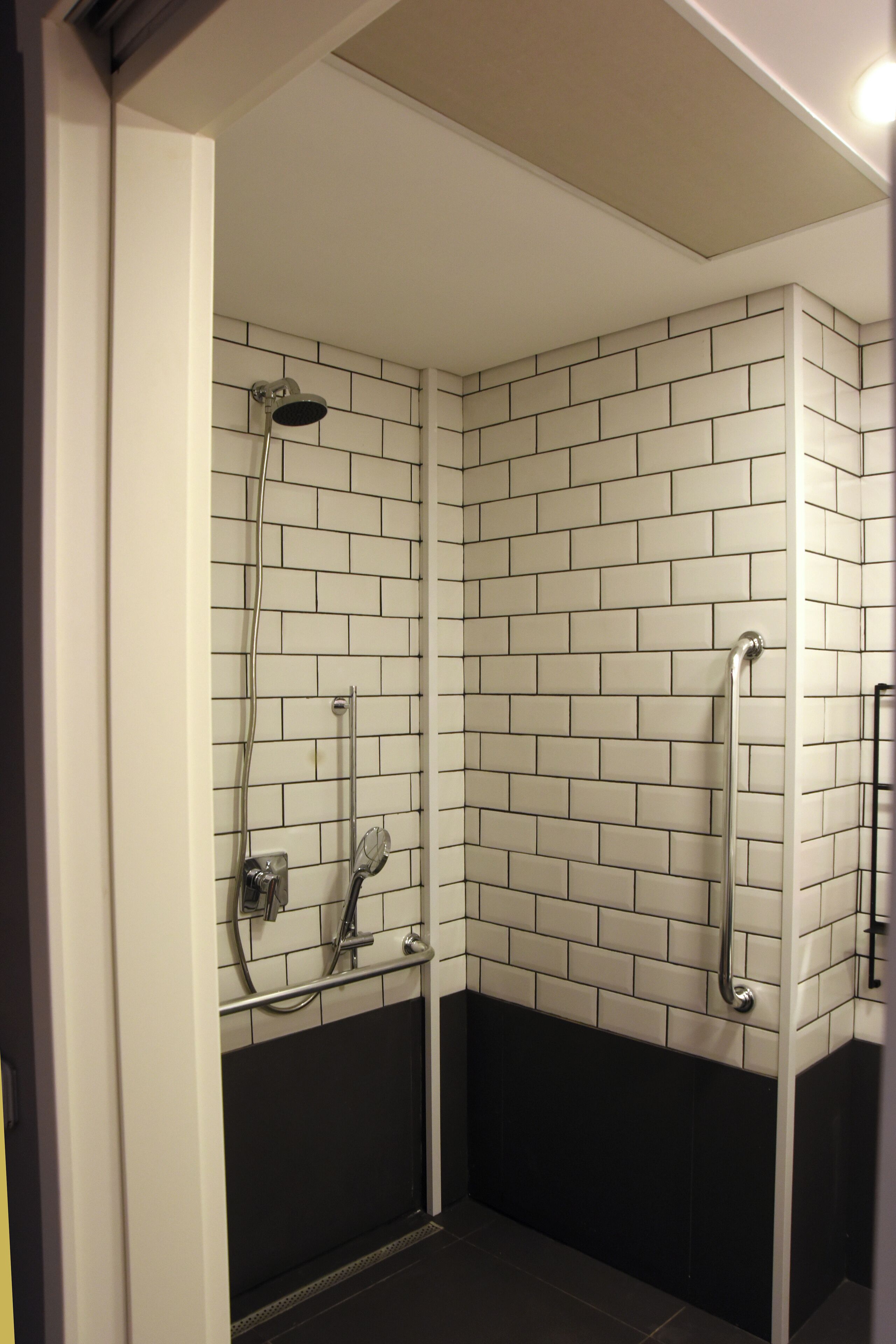 A small, enclosed shower area with white and black tiled walls, a handheld showerhead, and a vertical grab bar.