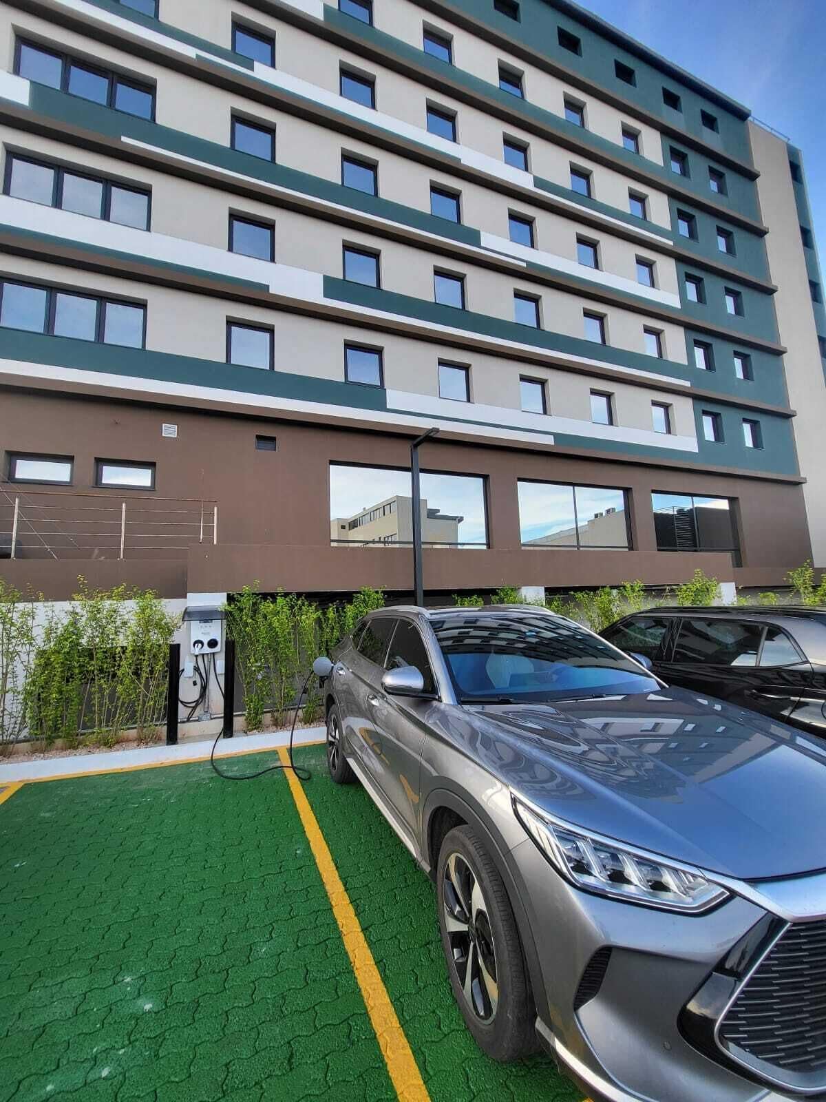 A modern building with multiple floors, a parking area with electric vehicle charging stations, and a green artificial turf surface.