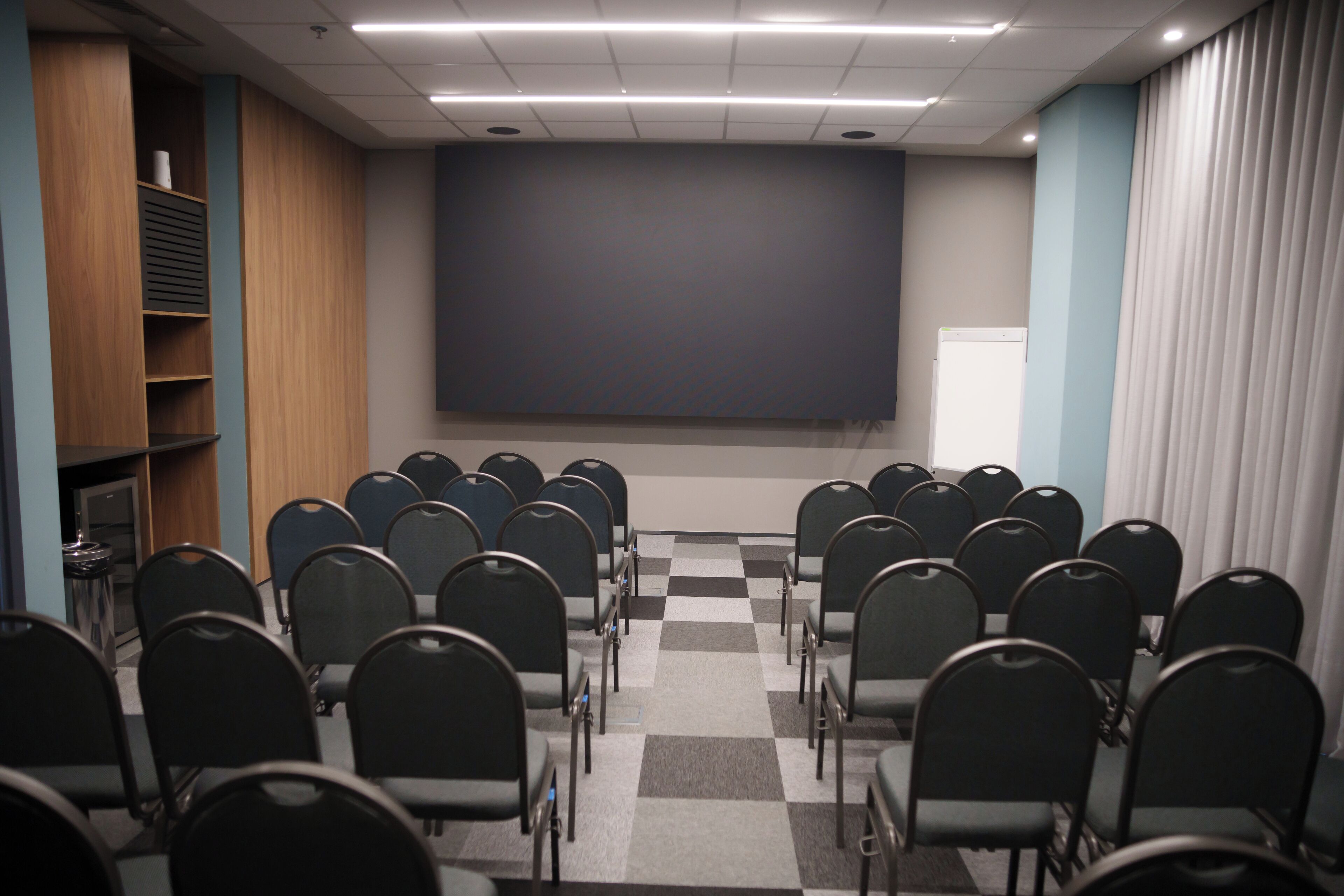 Un salón de conferencias con filas de sillas mirando hacia una pantalla grande, un armario de madera con estantes y un suelo a cuadros.