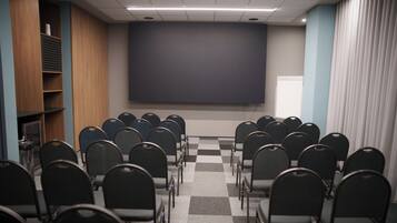 Un salón de conferencias con filas de sillas mirando hacia una pantalla grande, un armario de madera con estantes y un suelo a cuadros.