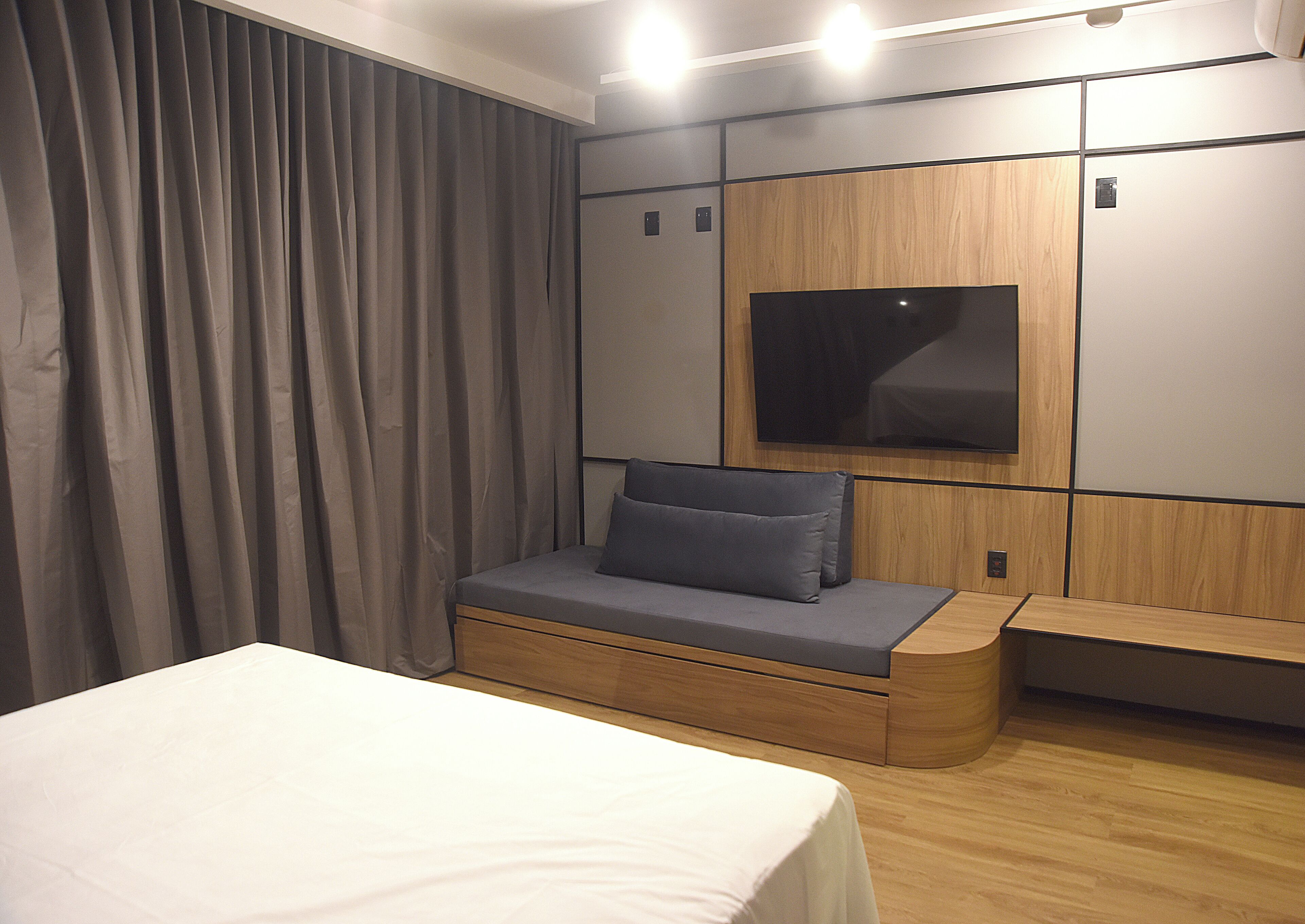Una habitación de hotel moderna con cama, una zona de estar con cojines, un televisor de pantalla plana montado en la pared y un diseño de paneles de madera.