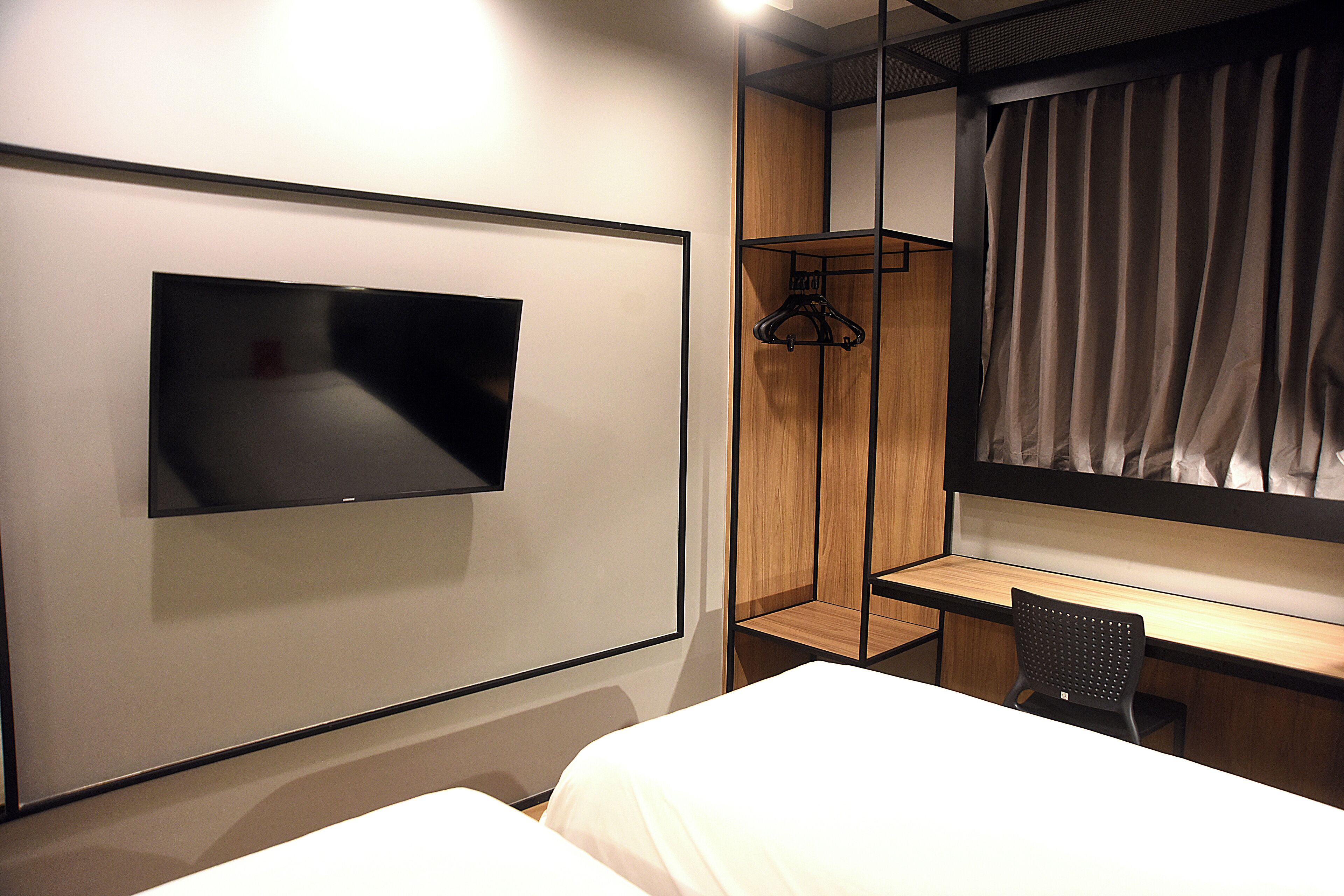 A modern hotel room with a flat-screen TV mounted on the wall, a wooden desk, a chair, and a bed with white bedding.