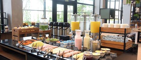 A buffet with various food items and beverages, including juices and milkshakes, in a well-lit room with large windows.