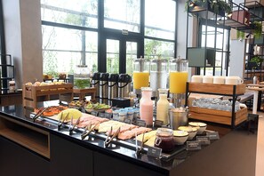 Un buffet proposant une variété de plats et de boissons, notamment des jus de fruits et des milkshakes, dans une pièce bien éclairée et dotée de grandes fenêtres.