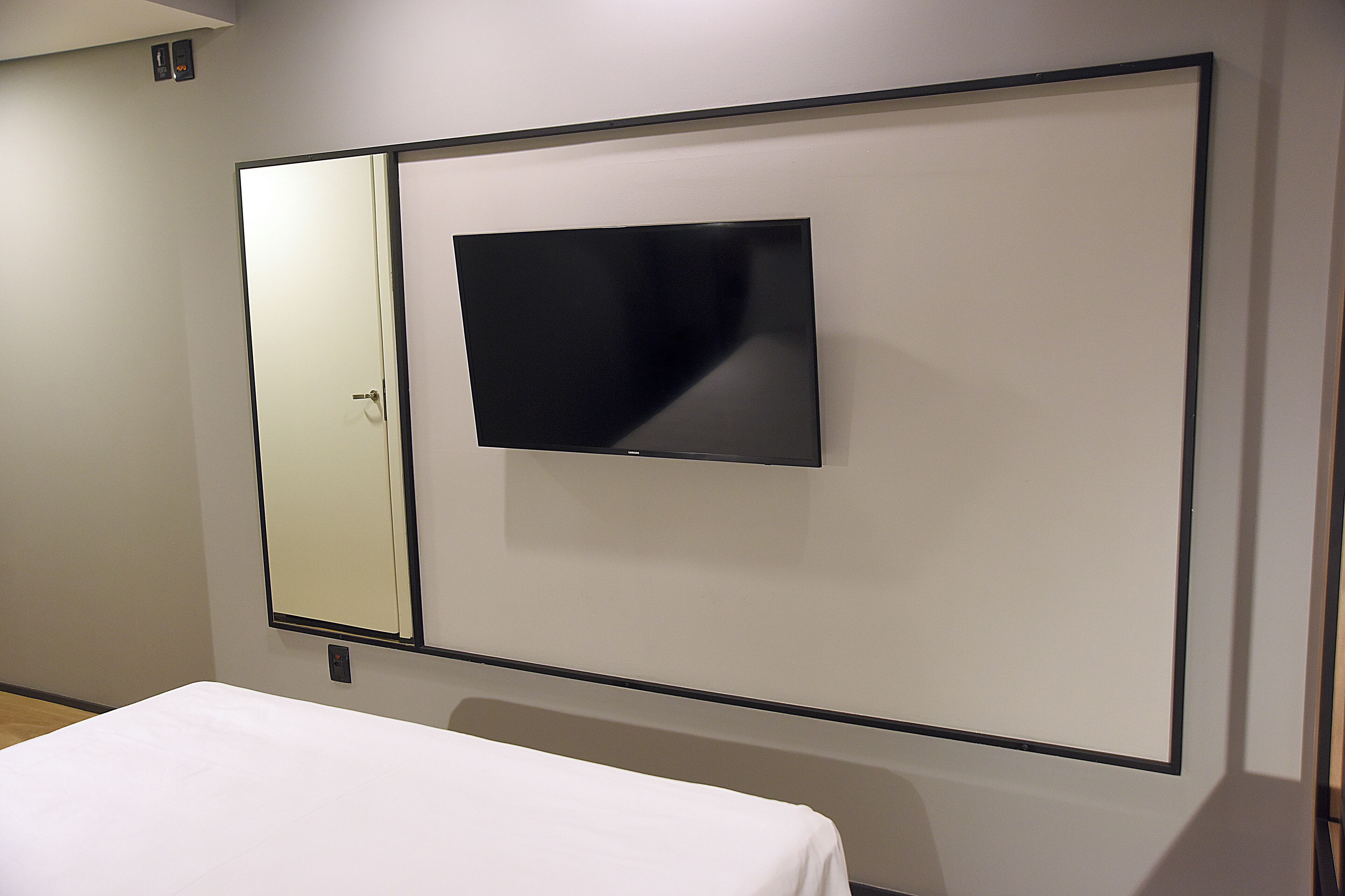 Una habitación de hotel con cama, un espejo grande y un televisor de pantalla plana montado en la pared.