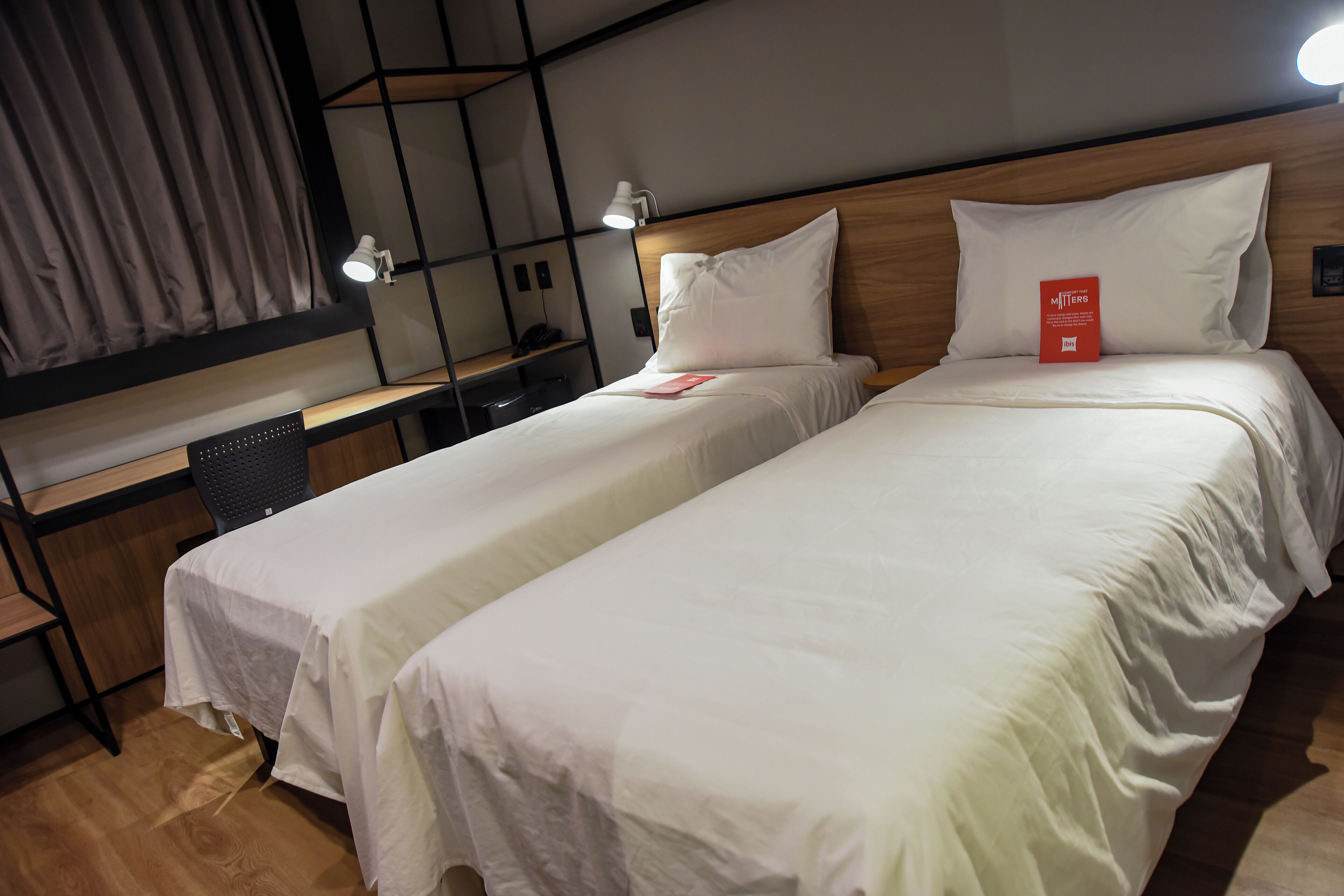 Een hotelkamer met twee eenpersoonsbedden, een bureau met stoel, een lamp en een rood boekje op het bed.