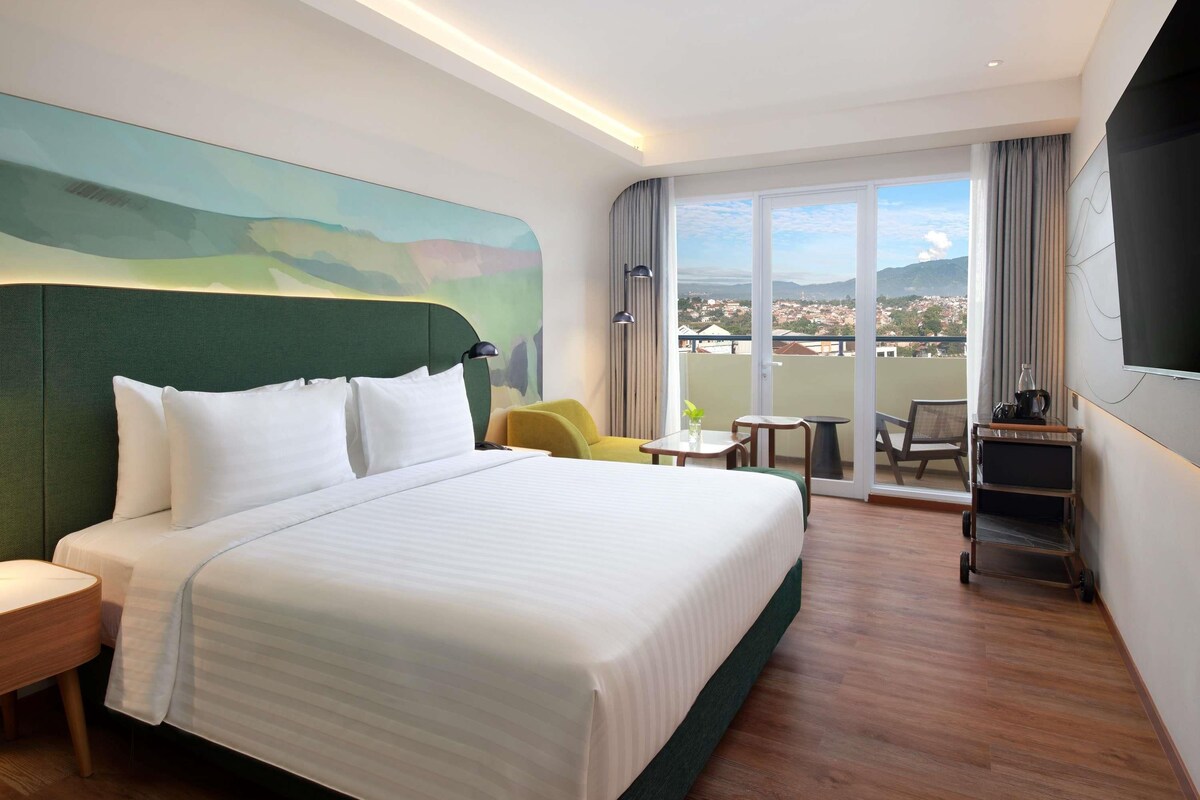 A hotel room with a large bed, a small table, a chair, a TV, and a balcony with a view.